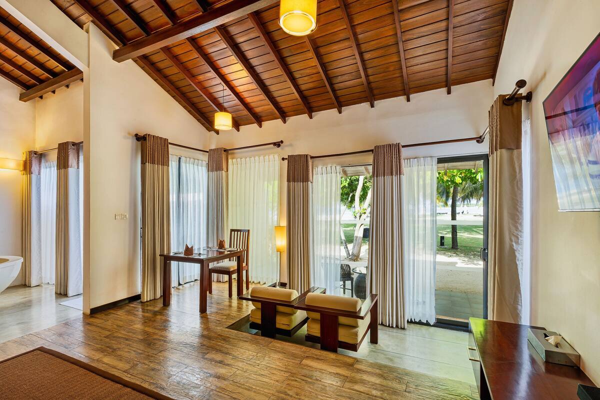 A spacious room with a wooden ceiling, a sofa, a table, and a view of a garden through a glass door.