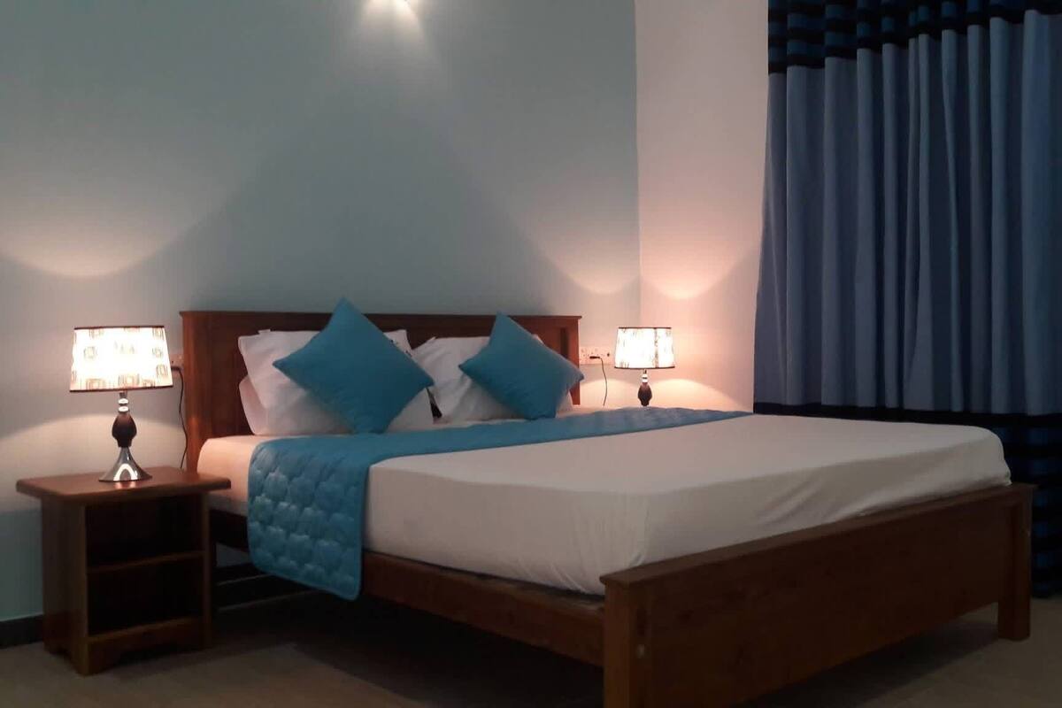 Una cama bien hecha con estructura de madera, dos mesitas de noche con lámparas y una cortina azul en la ventana.