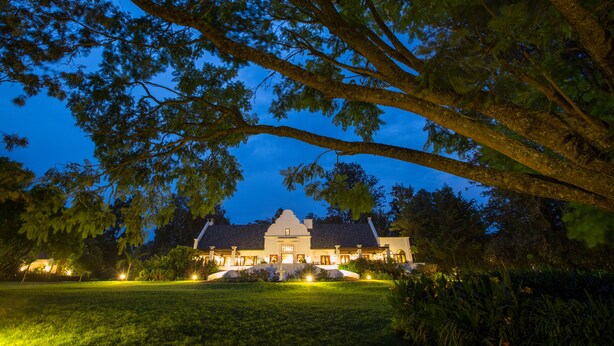 A large, well-lit building with a symmetrical facade, surrounded by lush greenery and a well-maintained lawn. - Elewana The Manor Ngorongoro (Karatu)