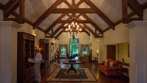 A spacious lobby with high ceilings and exposed wooden beams, a grand chandelier, a round table with a decorative centerpiece, and a red sofa. - Elewana The Manor Ngorongoro (Karatu)