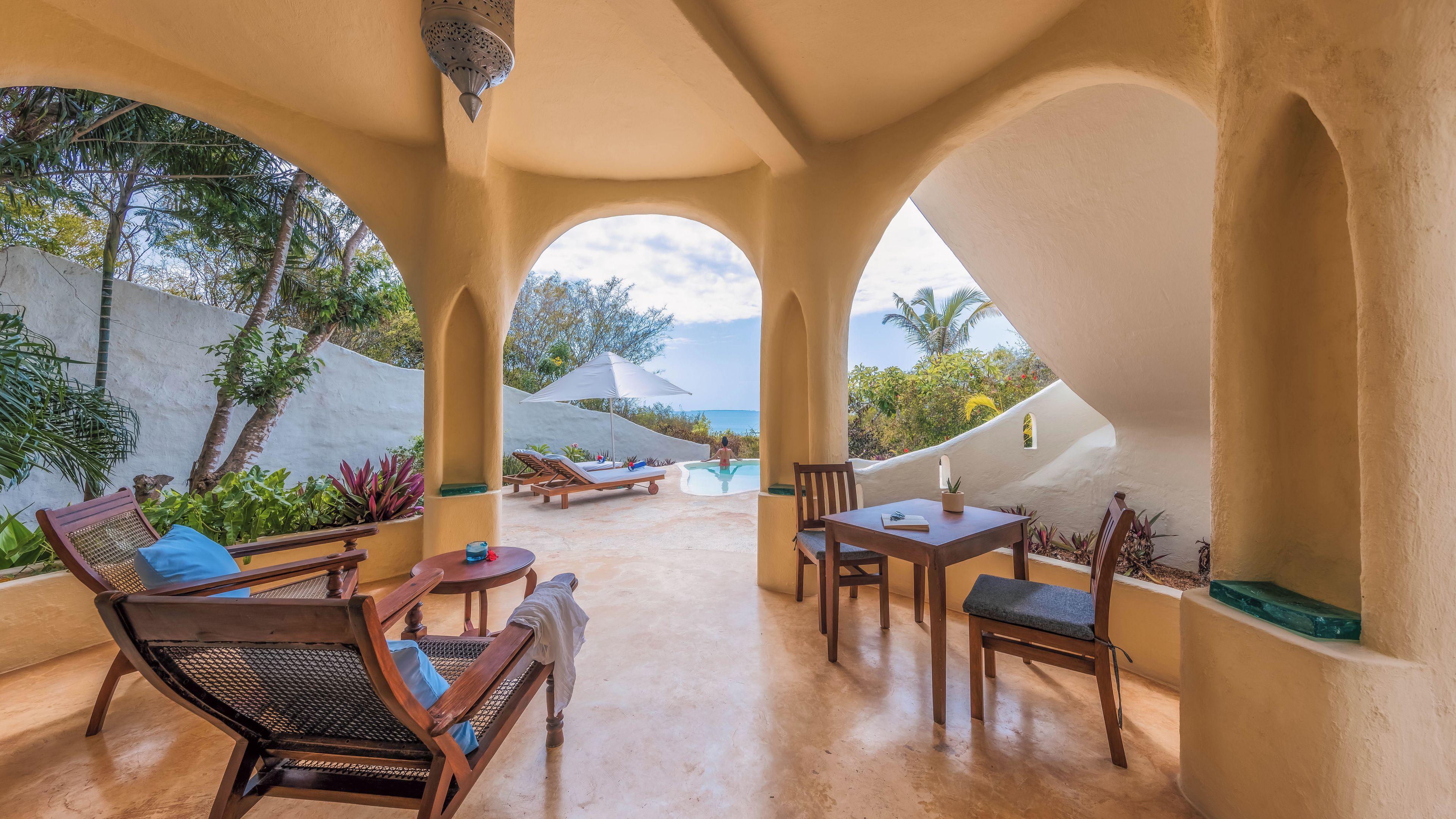 A sunlit outdoor lounge area with a pool, lounge chairs, and a table.