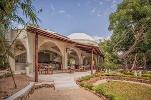 A covered outdoor seating area with tables and chairs, a stone pathway, and a garden with plants and trees. - Elewana Kilindi Zanzibar - Adults Only (Kendwa)