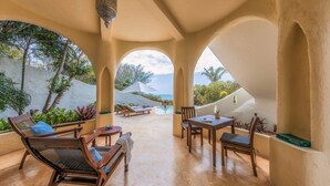 Un área de estar al aire libre techada con sillones, una mesa y vista a una piscina y al océano.