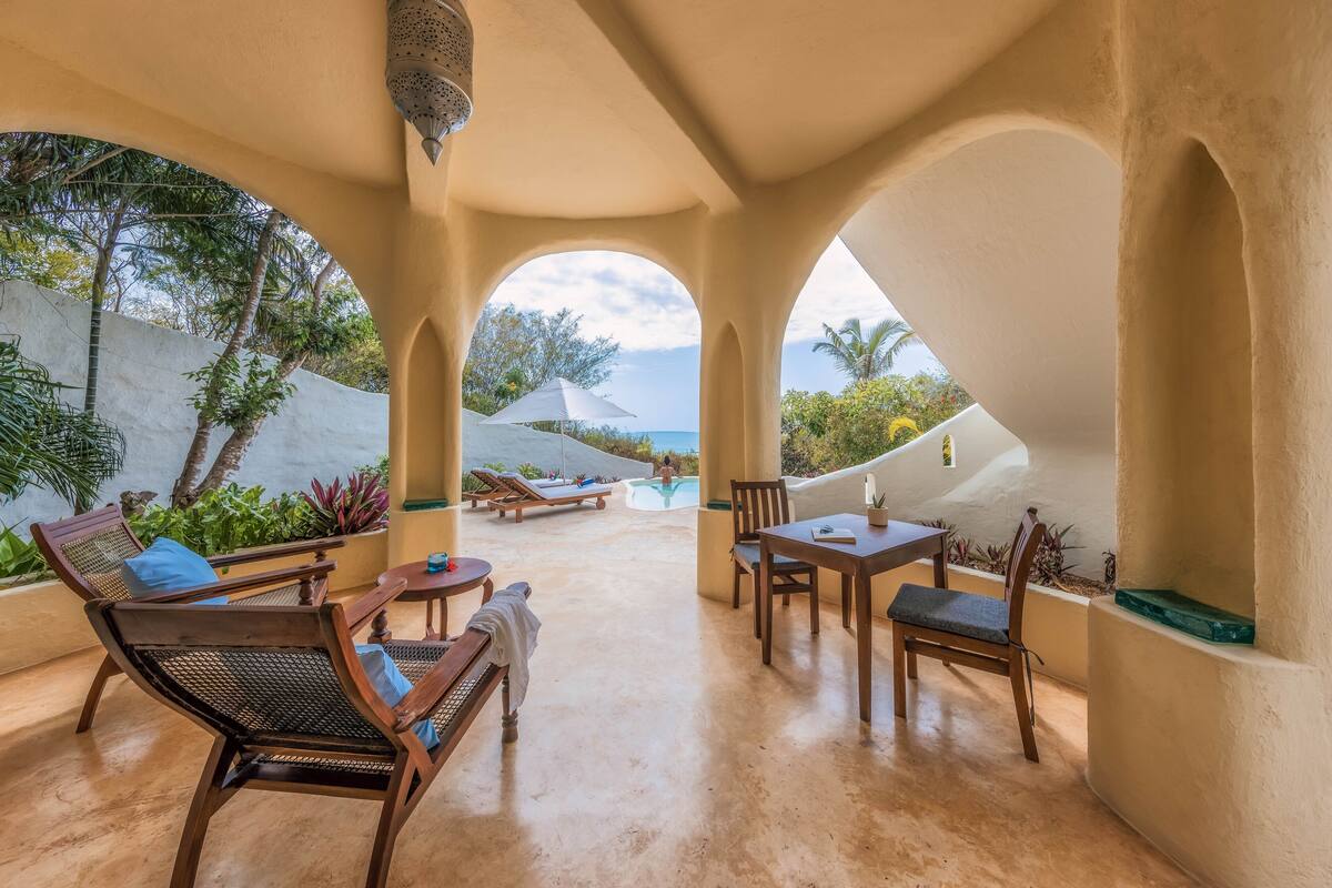A covered outdoor seating area with lounge chairs, a table, and a view of a pool and ocean.