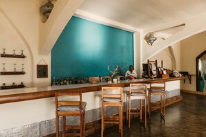 En bardisk med en turkosfärgad accentvägg, trästolar och en bartender.