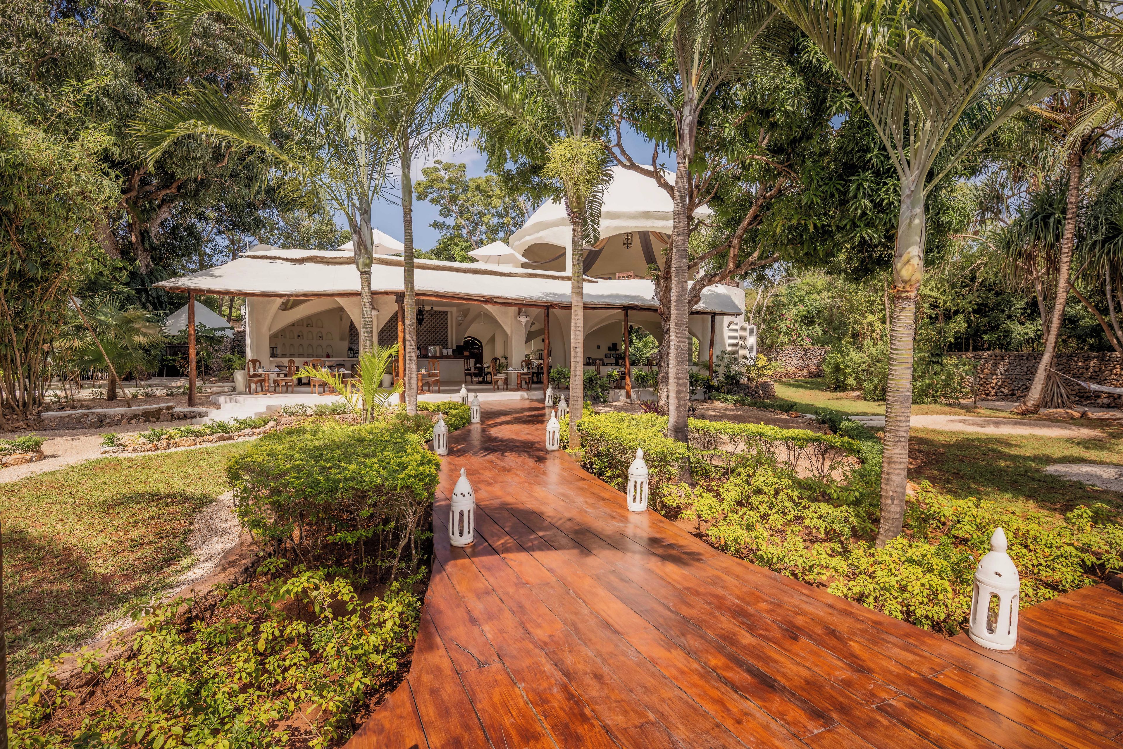 A resort with a covered outdoor seating area, a wooden pathway, and lush greenery.
