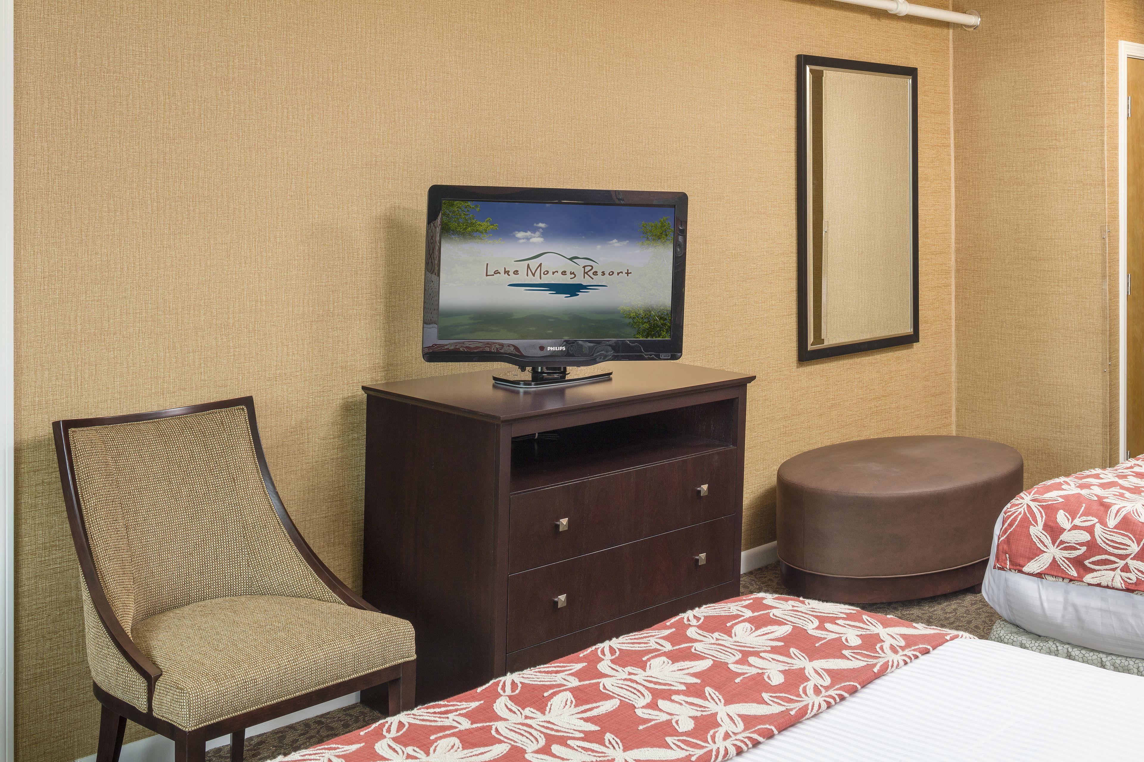 A hotel room with a television displaying 'Lake Morey Resort', a wooden chair, a brown ottoman, and a bed with a floral bedspread.