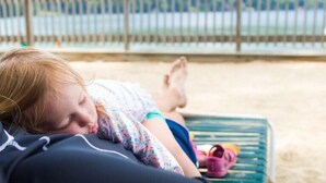 A person lying on a green lounge chair on a sandy beach with a view of a wooden fence and green hills in the background. - Lake Morey Resort (Fairlee)
