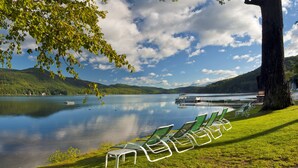 A lakeside view with a row of green lounge chairs under a tree, a dock with boats, and a calm lake reflecting the sky. - Lake Morey Resort (Fairlee)