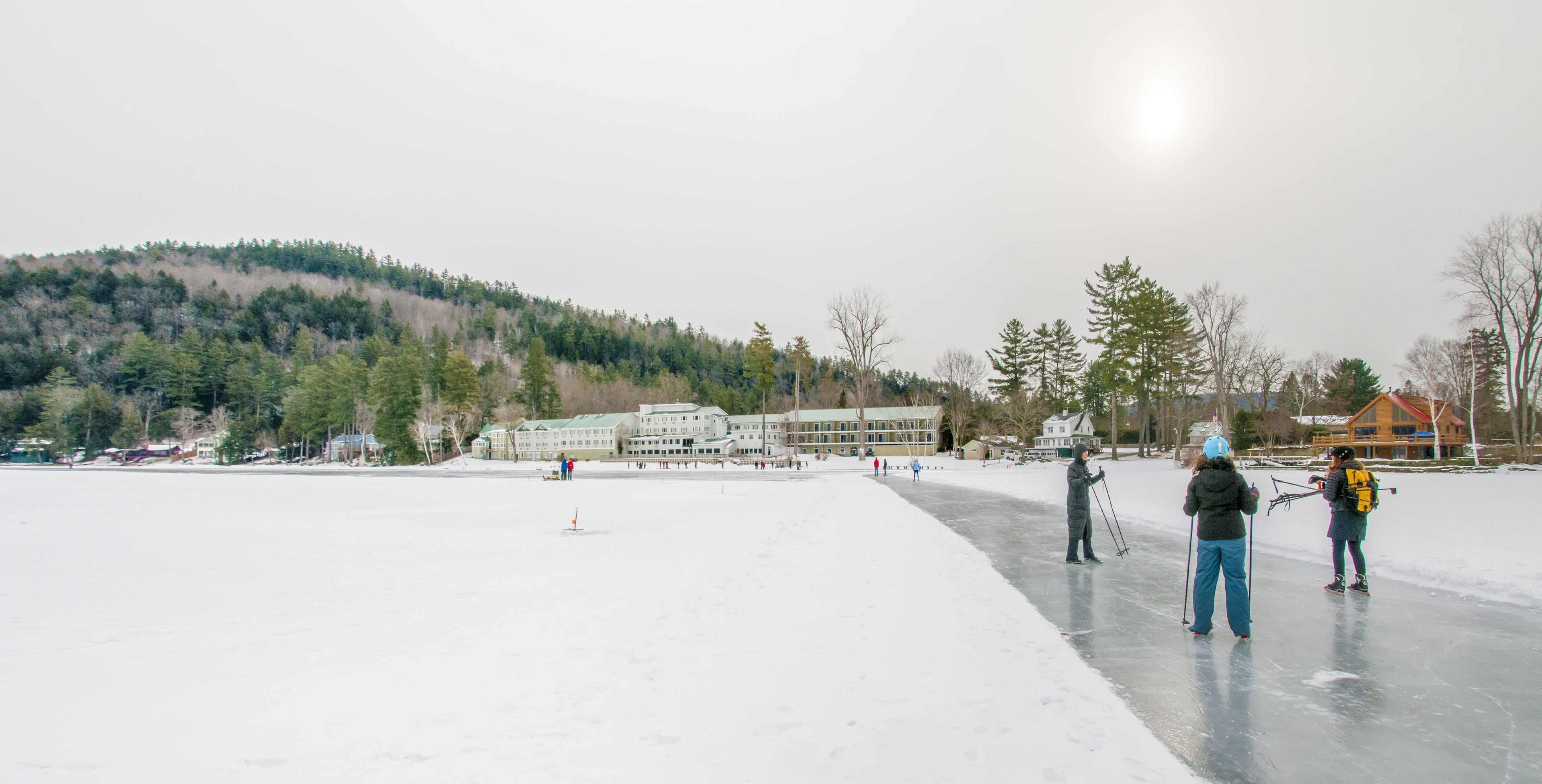 A frozen lake with people ice skating and skiing, surrounded by buildings and trees.