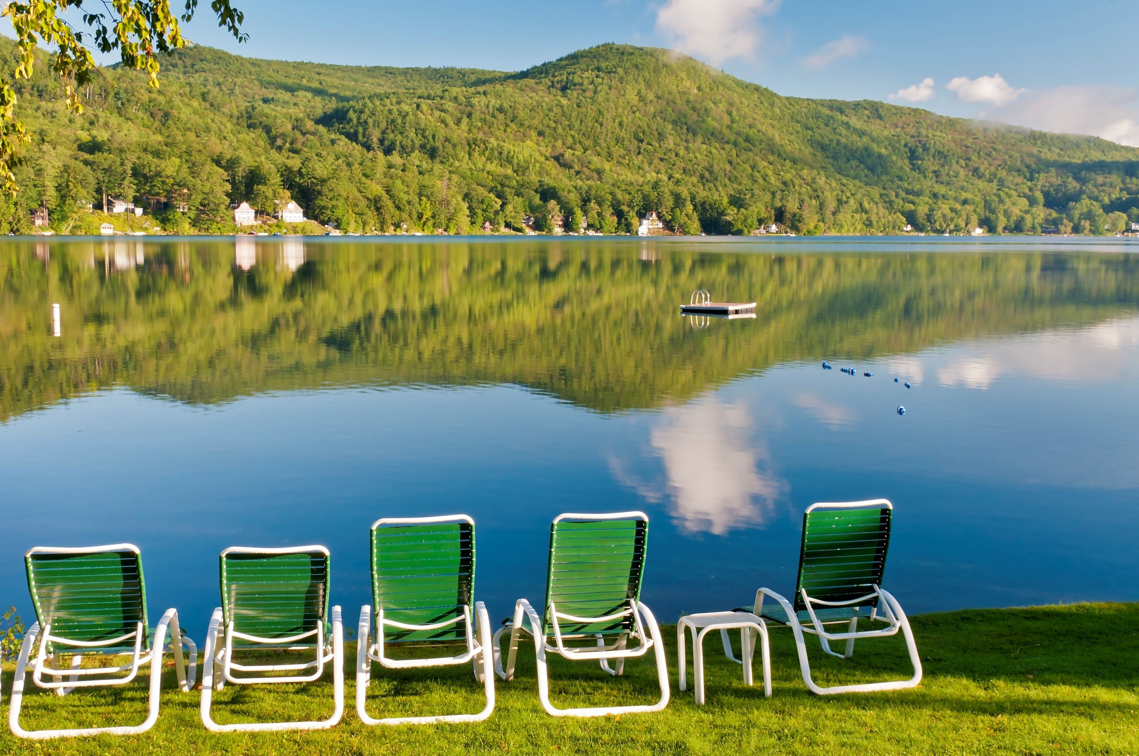 A serene lakeside view with a row of green chairs facing the water, a boat on the lake, and a backdrop of lush green hills.