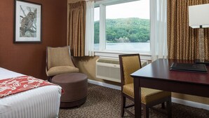 A hotel room with a bed, a desk, two chairs, a window with a view of a lake and forest, and an air conditioning unit. - Lake Morey Resort (Fairlee)