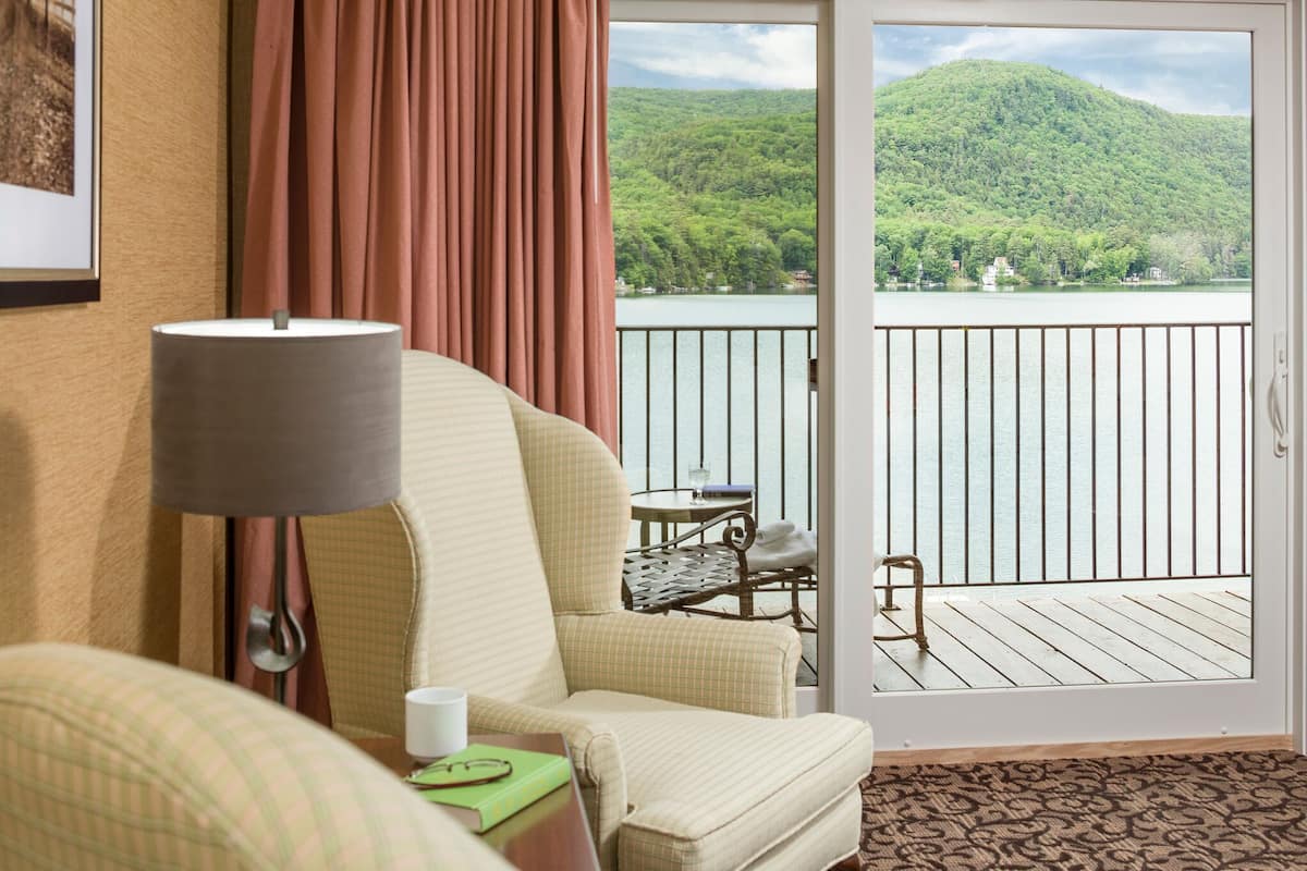 Ein Hotelzimmer mit einem karierten Sessel, einem Tisch mit Lampe und Blick auf einen See und Berge durch eine Glastür.