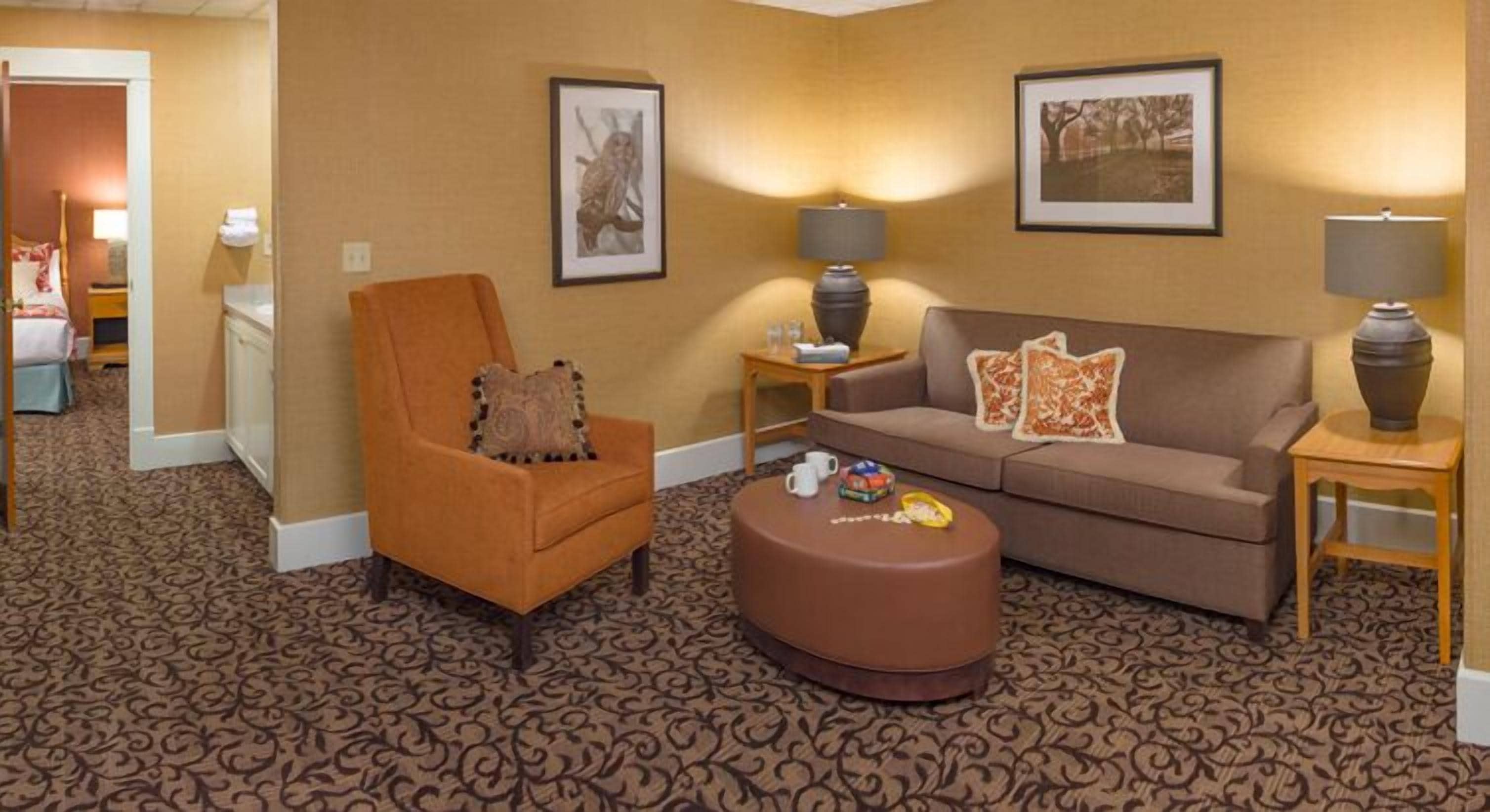 A hotel room with a patterned carpet, a brown armchair, a brown sofa, a small ottoman, and two side tables with lamps.