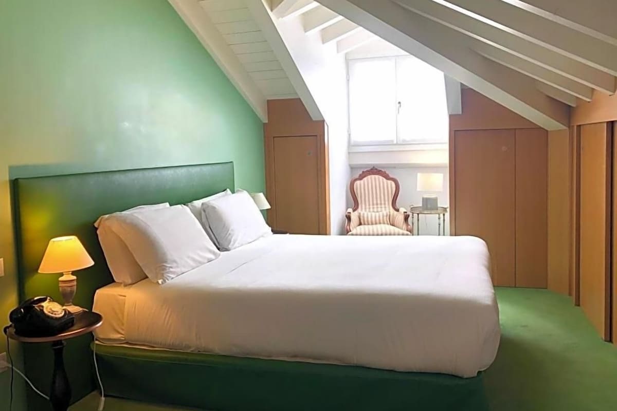 A single bed with a headboard, a bedside lamp, and a chair in a room with green walls and a sloped ceiling.