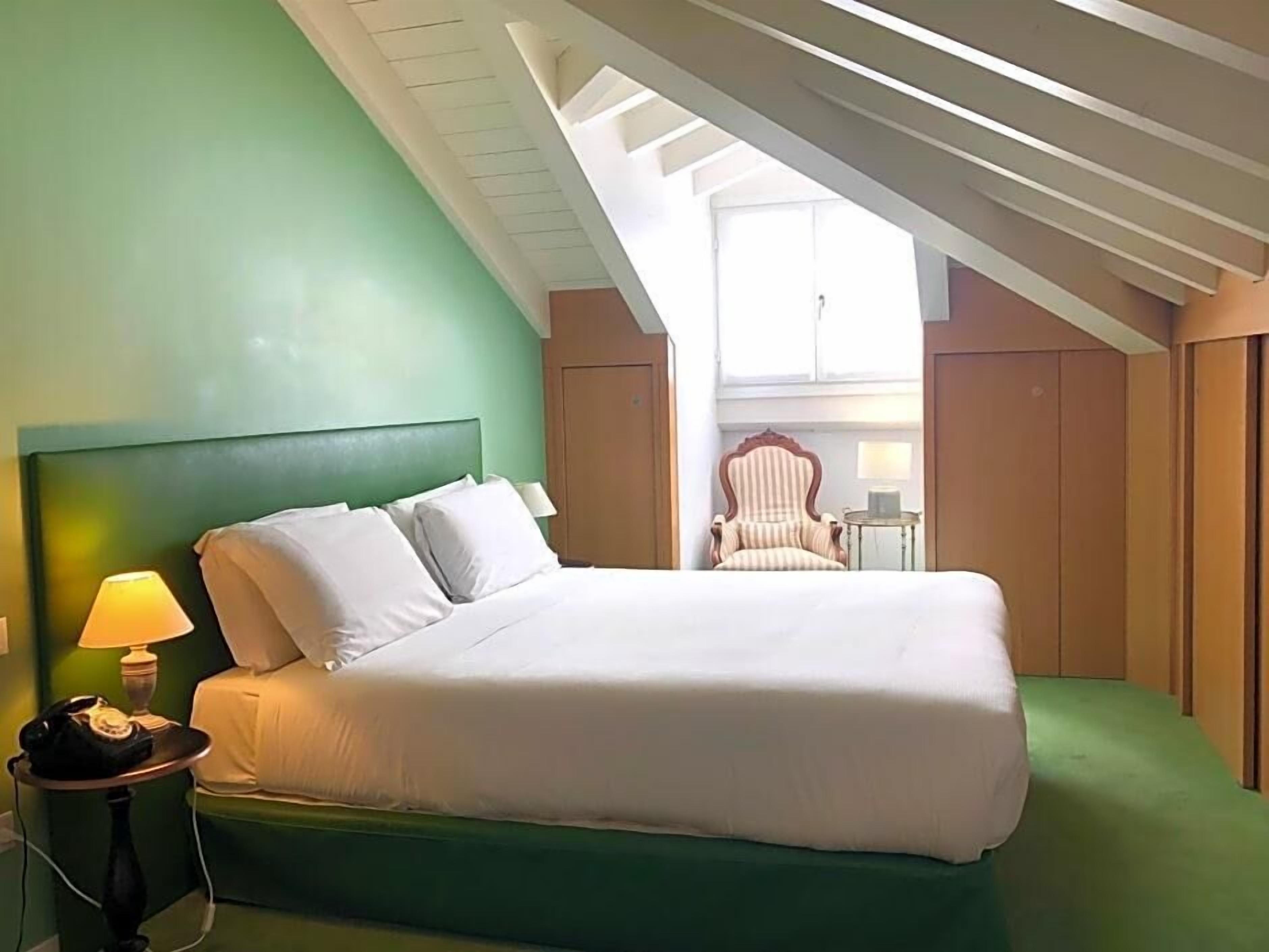 A single bed with a headboard, a bedside lamp, and a chair in a room with green walls and a sloped ceiling.