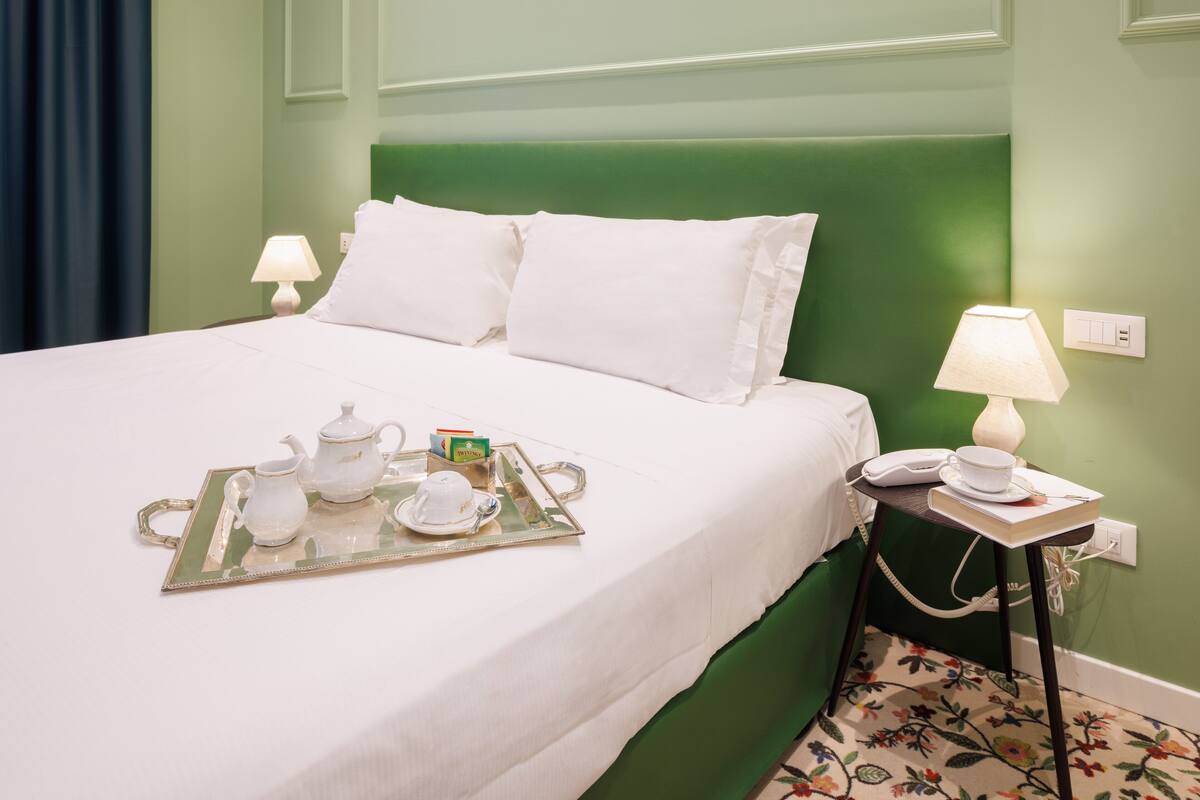 A neatly made bed with white linens and pillows, a tray with tea set, and a bedside table with a lamp and phone.