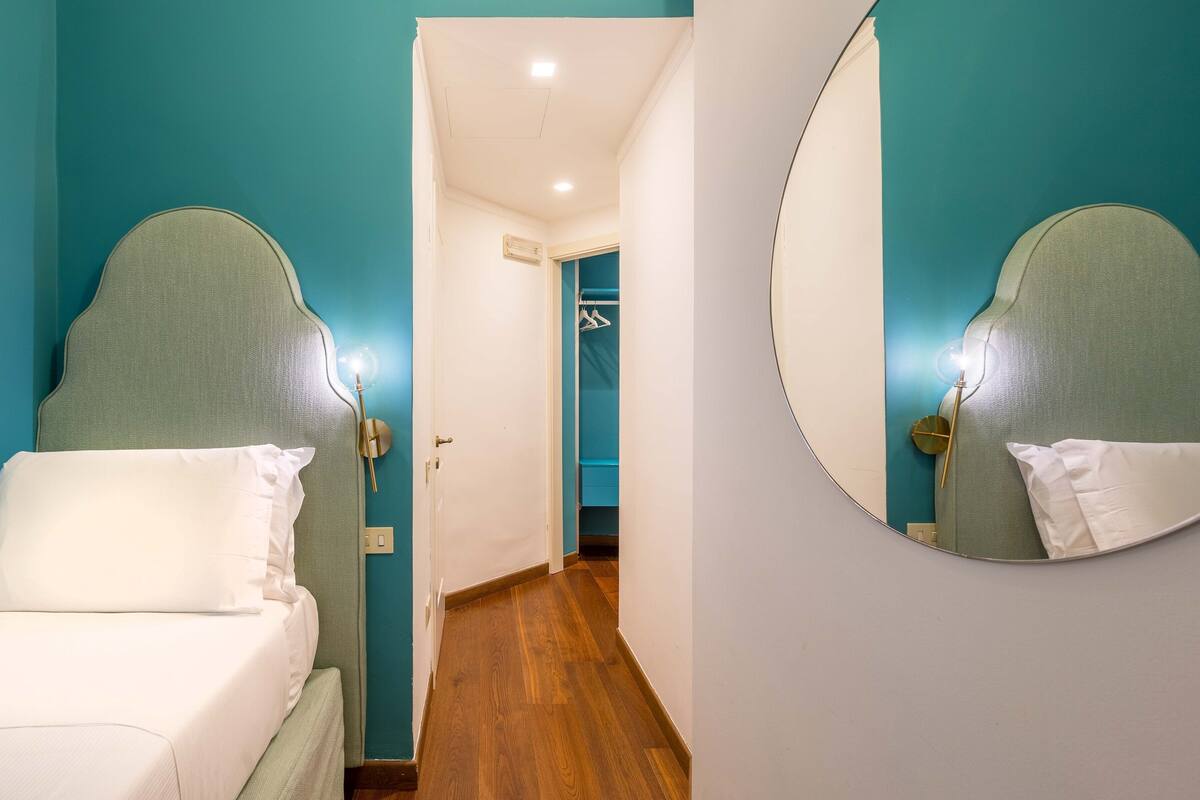 A hotel room with teal walls, a wooden floor, a bed with white bedding, a round mirror, and a hallway leading to a bathroom.