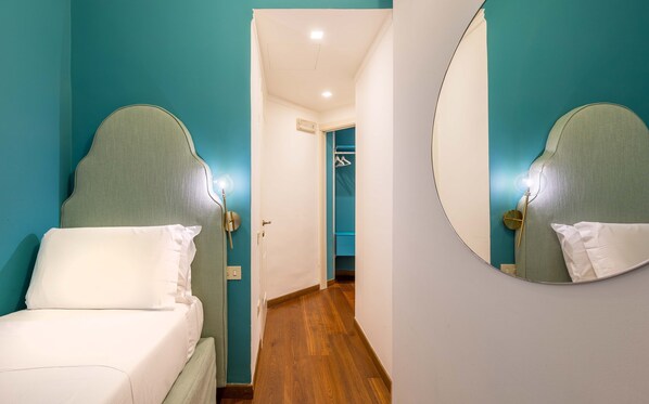 A hotel room with teal walls, a wooden floor, a bed with white bedding, a round mirror, and a hallway leading to a bathroom. - Antica Locanda Leonardo (Milan)
