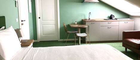 A compact room with a bed, a desk with a chair, a kitchenette, and a red chair.