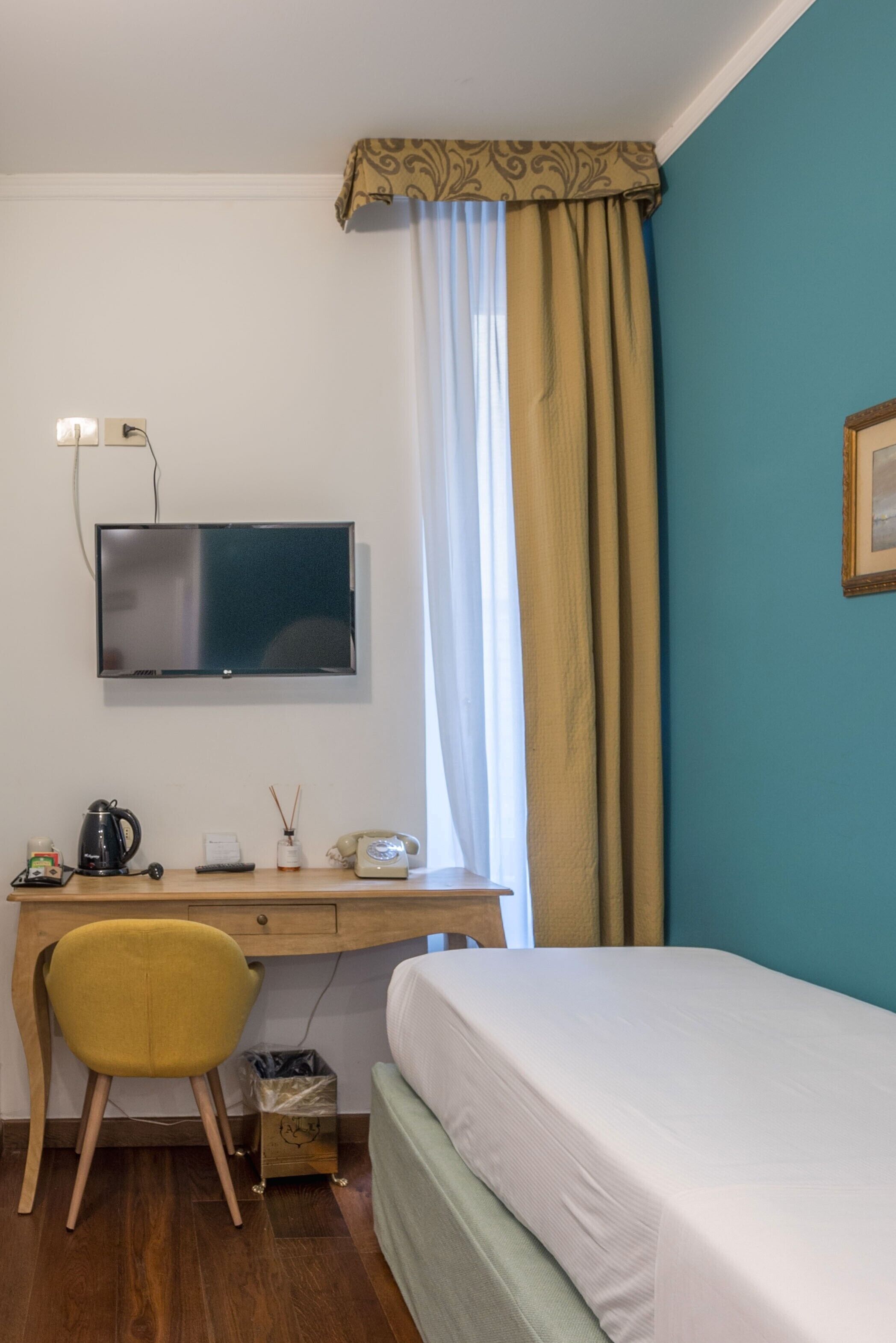 A room with a bed, a desk with a computer, a chair, a TV mounted on the wall, and a window with curtains.