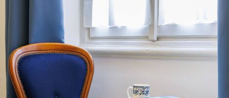 A room with a blue upholstered chair, a small round table with a book and a mug, and a window with curtains.