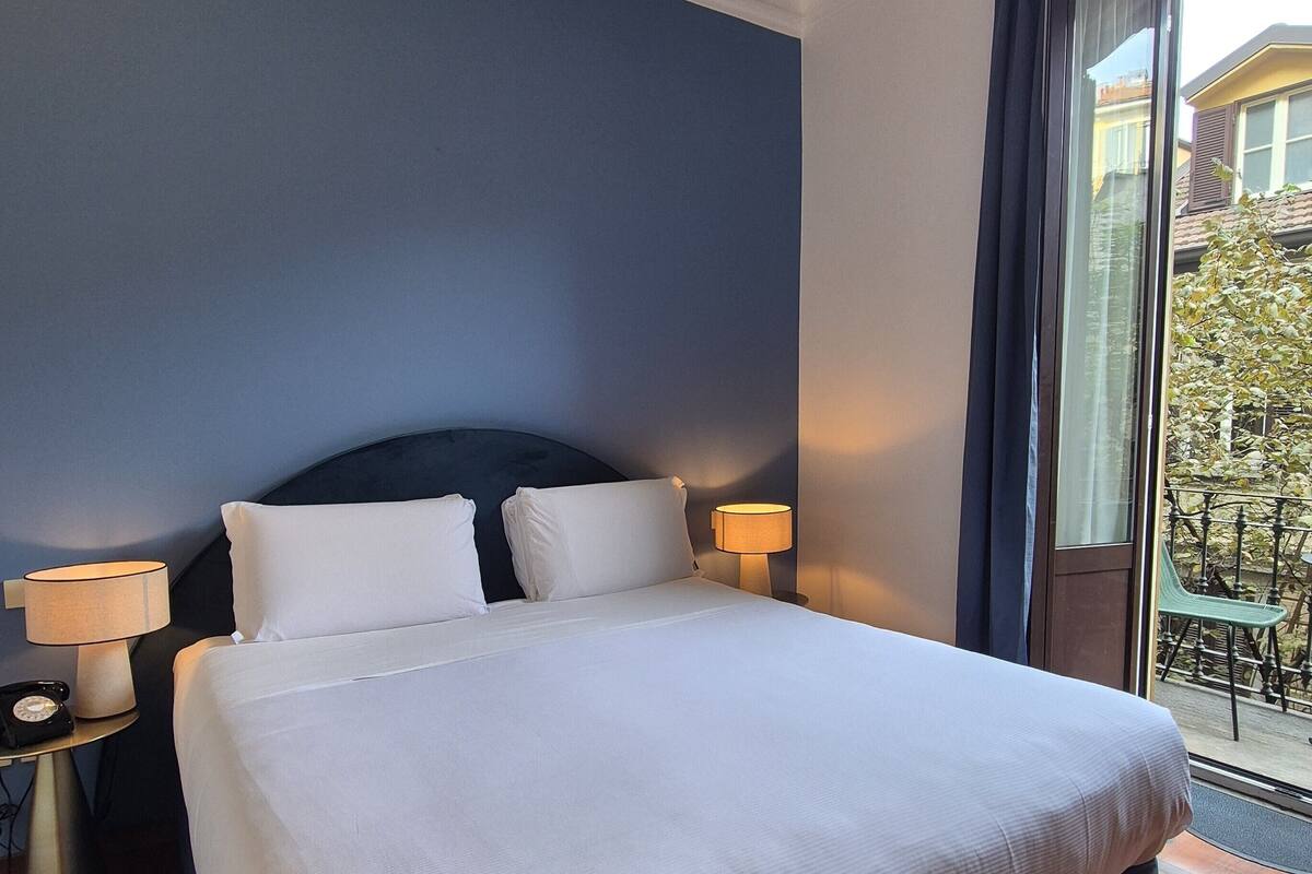 A hotel room with a bed, two bedside lamps, a telephone, and a window with a view of the outside.