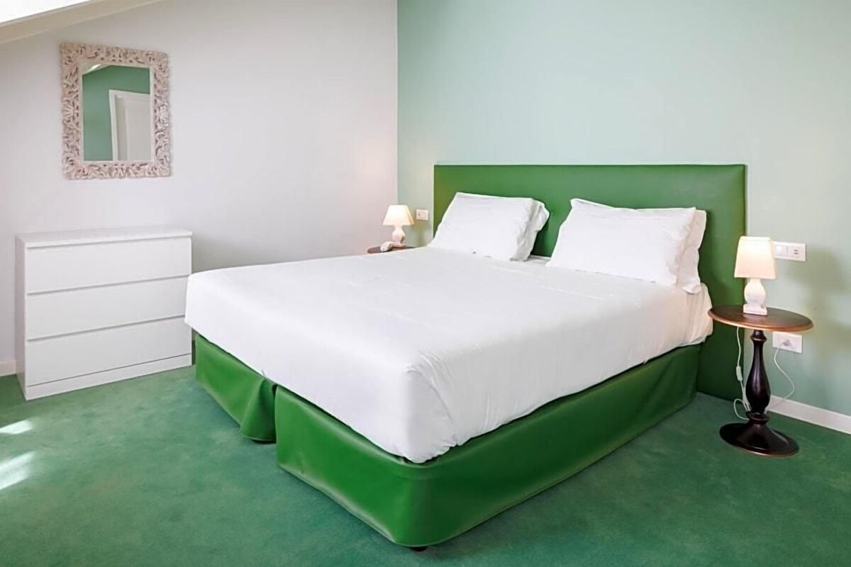 Ein Zimmer mit einem dunkelgrünen Bett, weißer Bettwäsche, einer weißen Kommode und zwei Nachttischlampen.