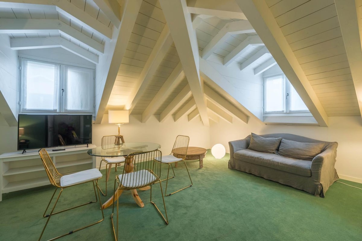 A loft-style room with a high ceiling, a dining area with a glass table and metal chairs, a sofa, and a television.