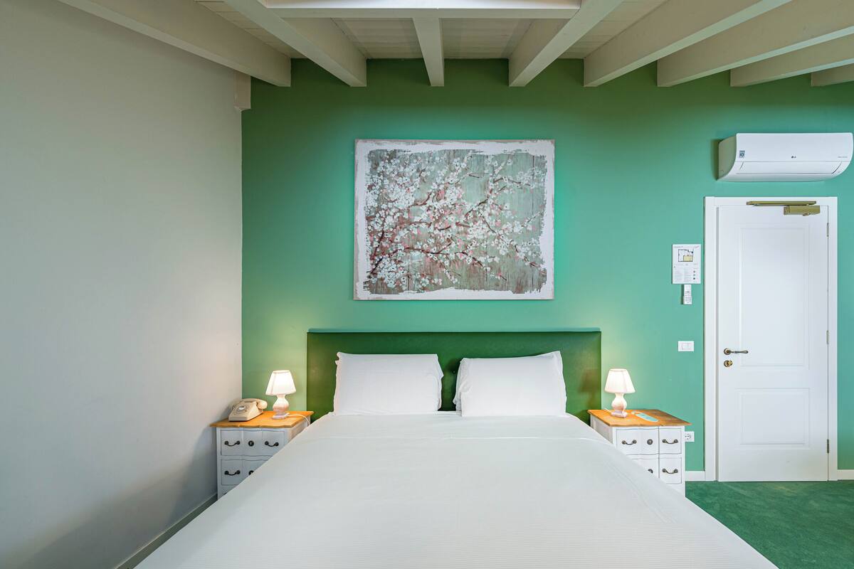 A bedroom with a bed, two bedside tables with lamps, a white door, and a green wall with a floral painting.
