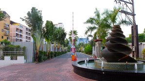 A paved pathway with a decorative fountain, palm trees, and buildings in the background.
