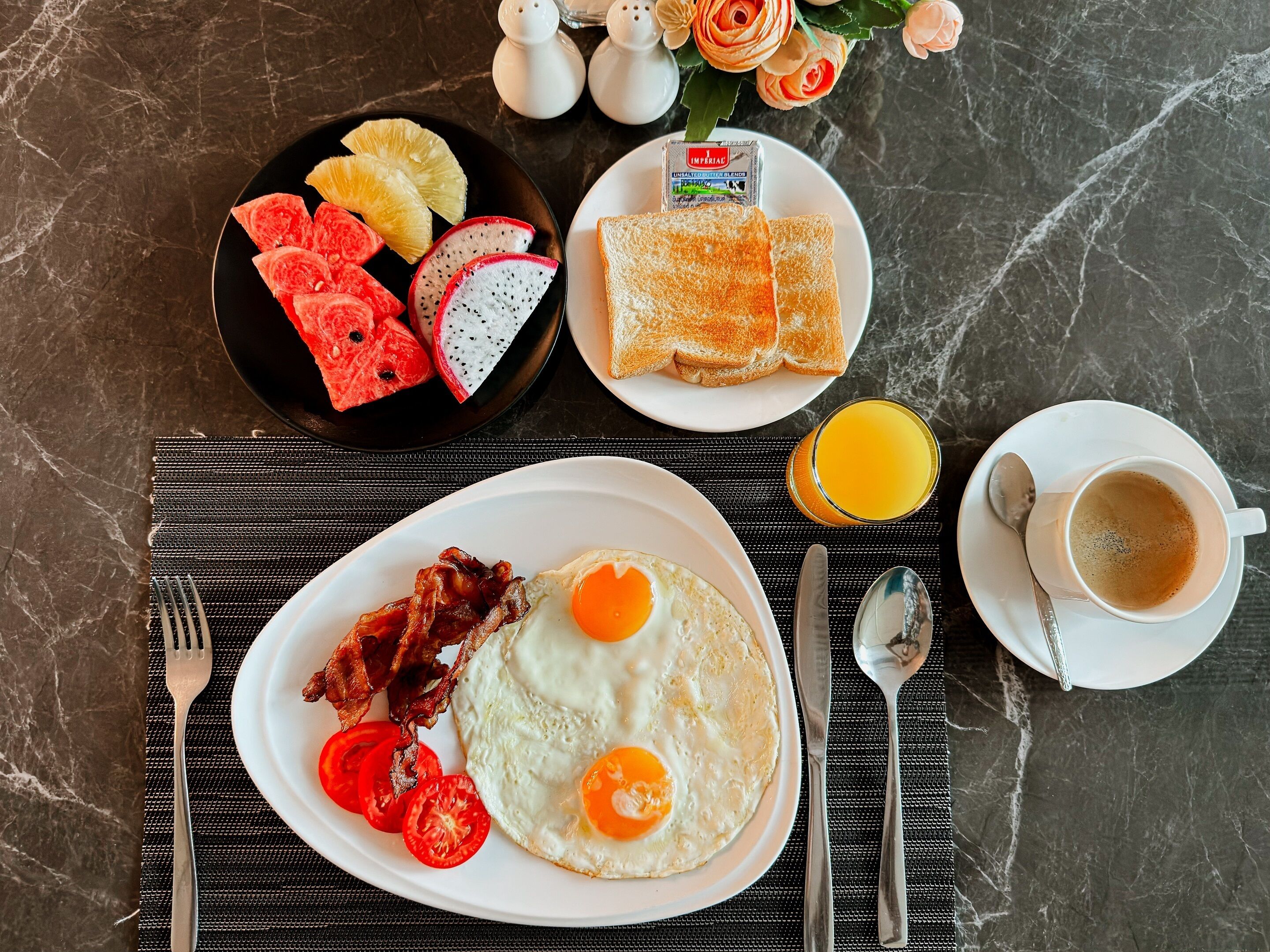 Une table en marbre dressée pour le petit-déjeuner, avec des toasts, des œufs, du bacon, des tomates et des fruits.