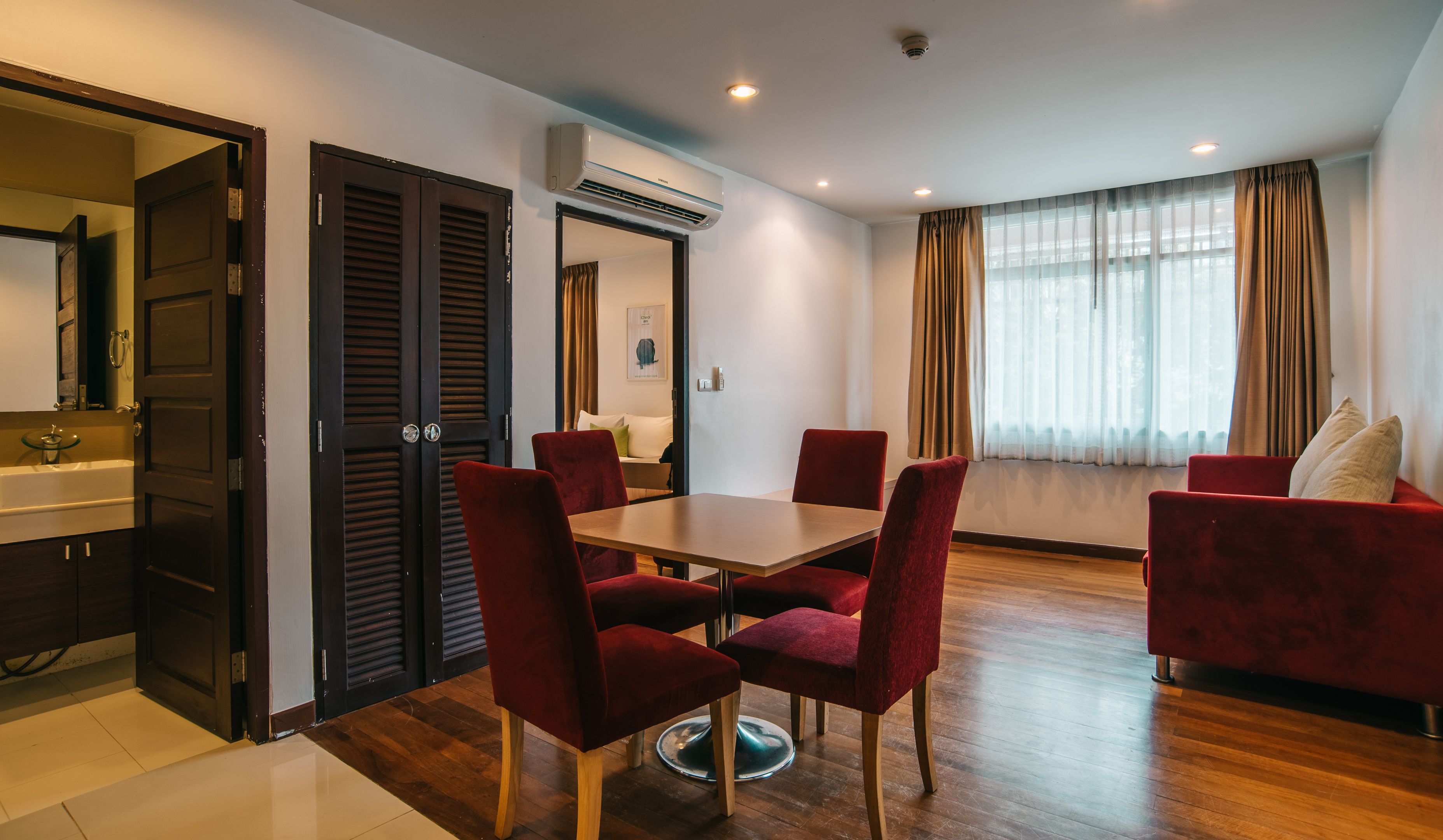 A modern hotel room with a dining area, a red sofa, a wooden table, and a large window with curtains.
