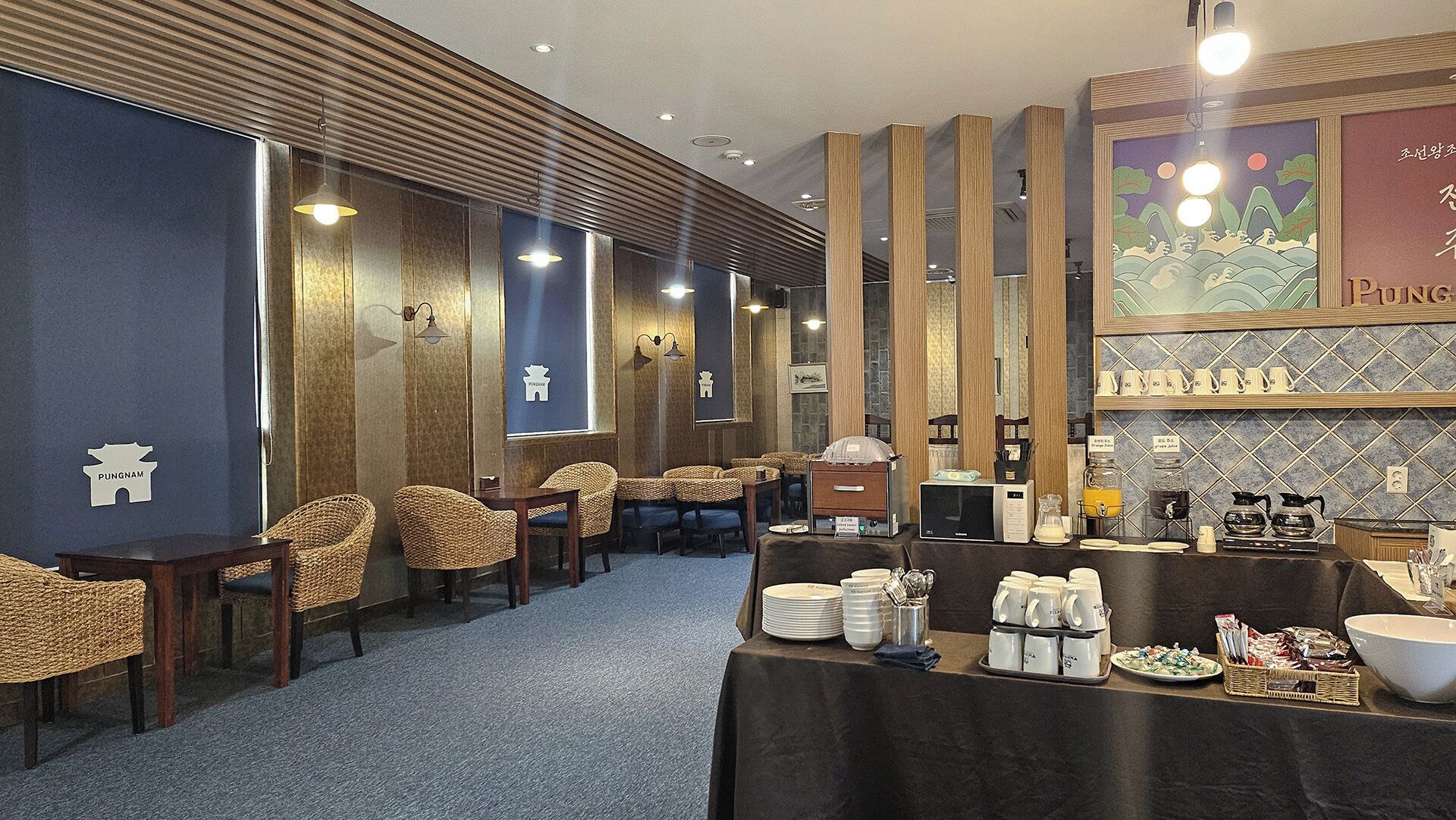 Un restaurant avec des chaises en osier, une table de buffet garnie de plats et un mur orné d’un tableau encadré.