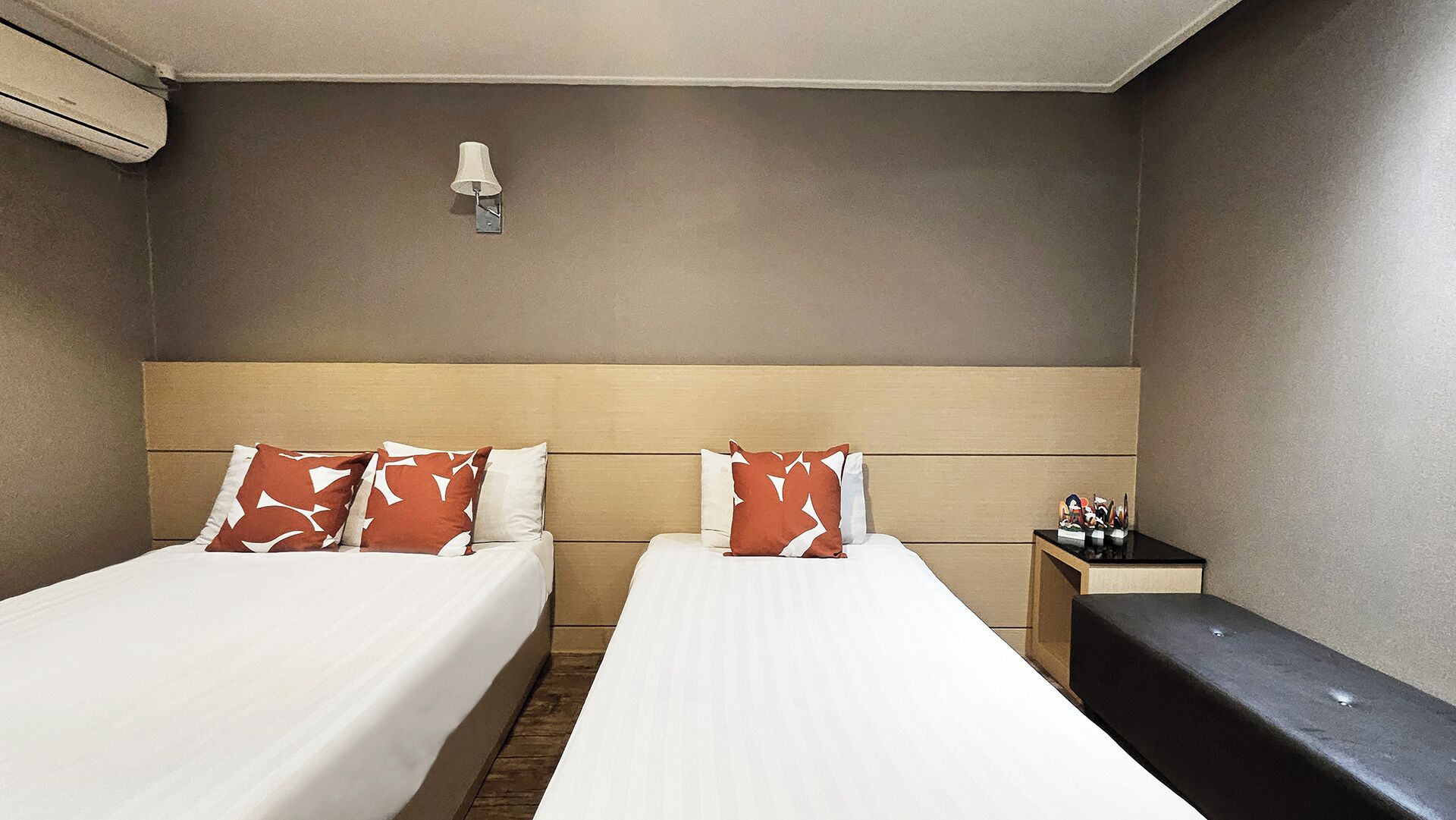 Quarto de hotel com duas camas, uma mesa de cabeceira, um banco e uma luminária fixada na parede.