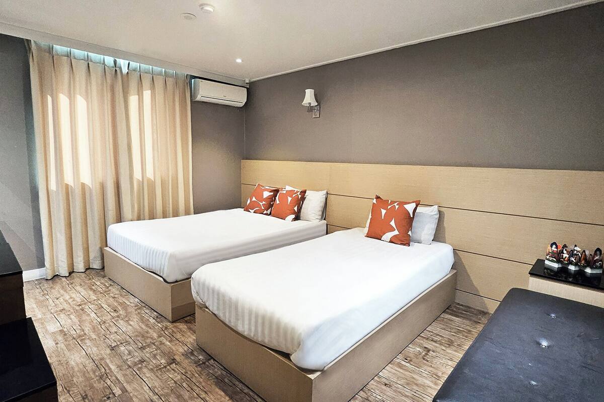 A hotel room with two beds, a desk, a chair, a lamp, and an air conditioning unit.