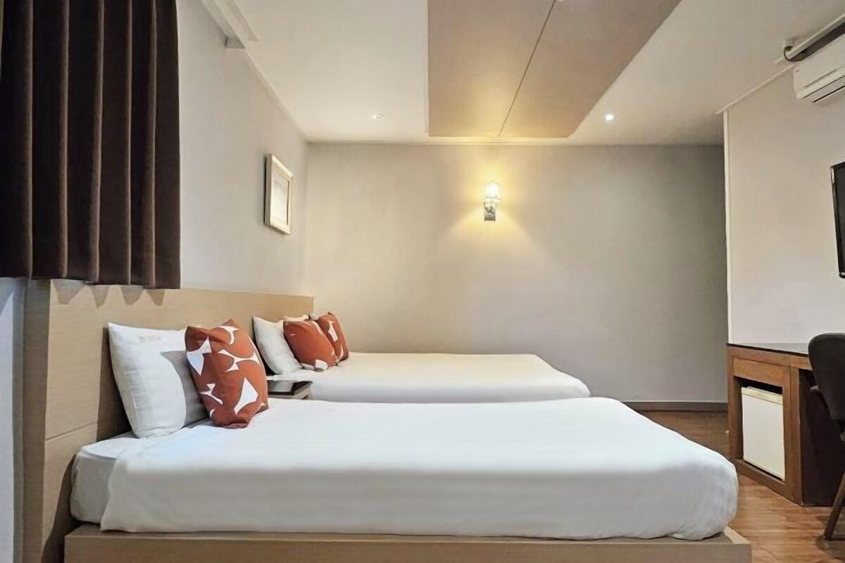 Ett hotellrum med två sängar, ett skrivbord, en stol, en väggmonterad lampa och ett fönster med persienner.