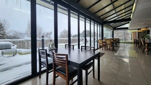 Une salle à manger spacieuse, dotée de grandes fenêtres, d’un haut plafond et offrant une vue sur une terrasse extérieure enneigée.