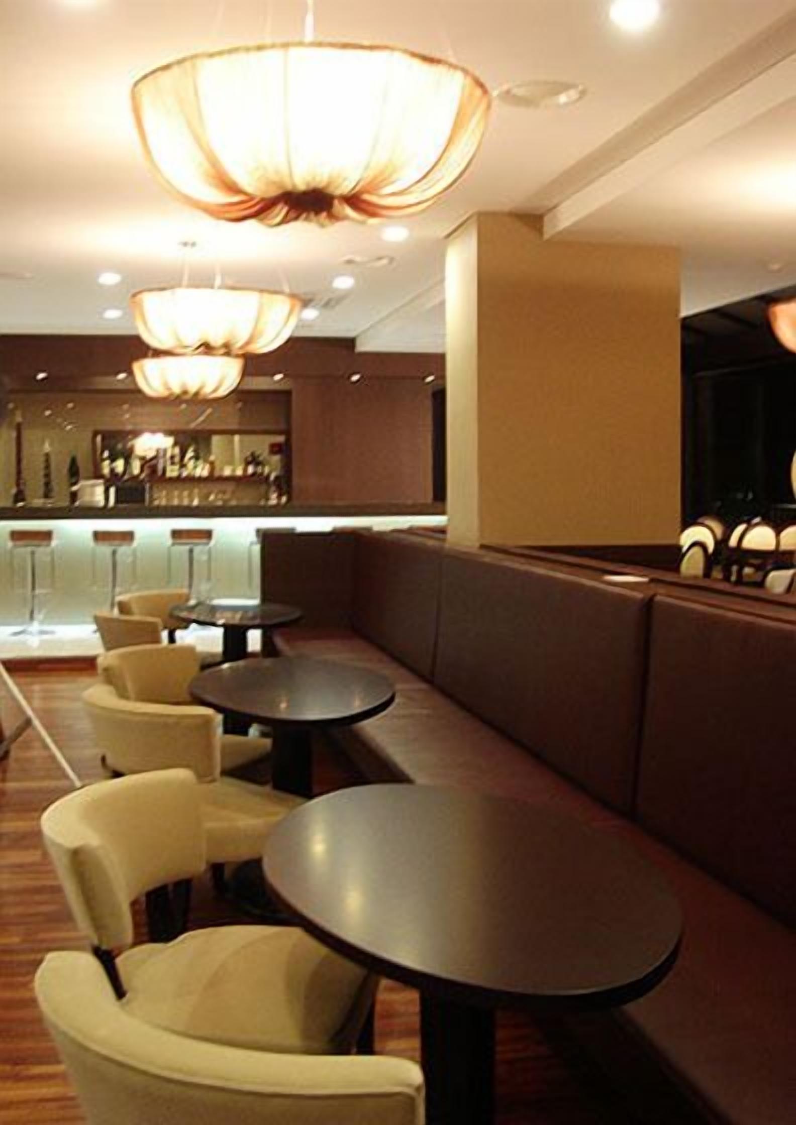 A restaurant with booths and round tables, overhead lighting, and a bar area in the background.
