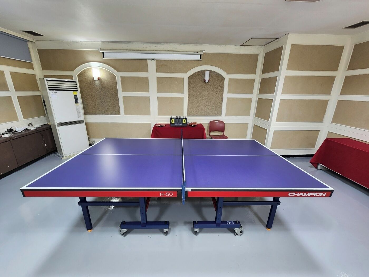 Une table de ping-pong portant la mention « H-50 » et la marque « CHAMPION », placée dans une pièce dont les murs sont recouverts de panneaux beiges et blancs, avec une table rouge et un réfrigérateur blanc.