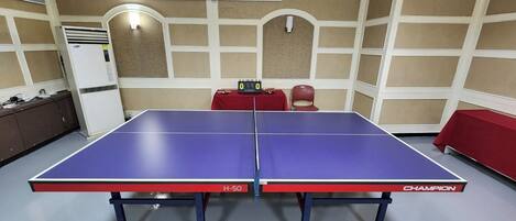 Une table de ping-pong portant la mention « H-50 » et la marque « CHAMPION », placée dans une pièce dont les murs sont recouverts de panneaux beiges et blancs, avec une table rouge et un réfrigérateur blanc.