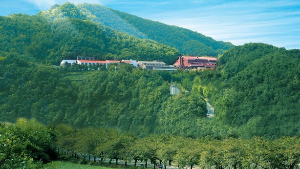 Un hôtel niché dans une vallée verdoyante, avec une chaîne de montagnes en toile de fond, se distinguant par un bâtiment rouge et une structure blanche surmontée d’un toit rouge.