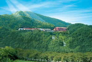 Un hôtel niché dans une vallée verdoyante, avec une chaîne de montagnes en toile de fond, se distinguant par un bâtiment rouge et une structure blanche surmontée d’un toit rouge.