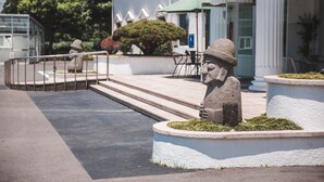 Una estatua de un hombre con sombrero sobre una plataforma circular rodeada de césped, frente a un edificio con columnas y un toldo verde.