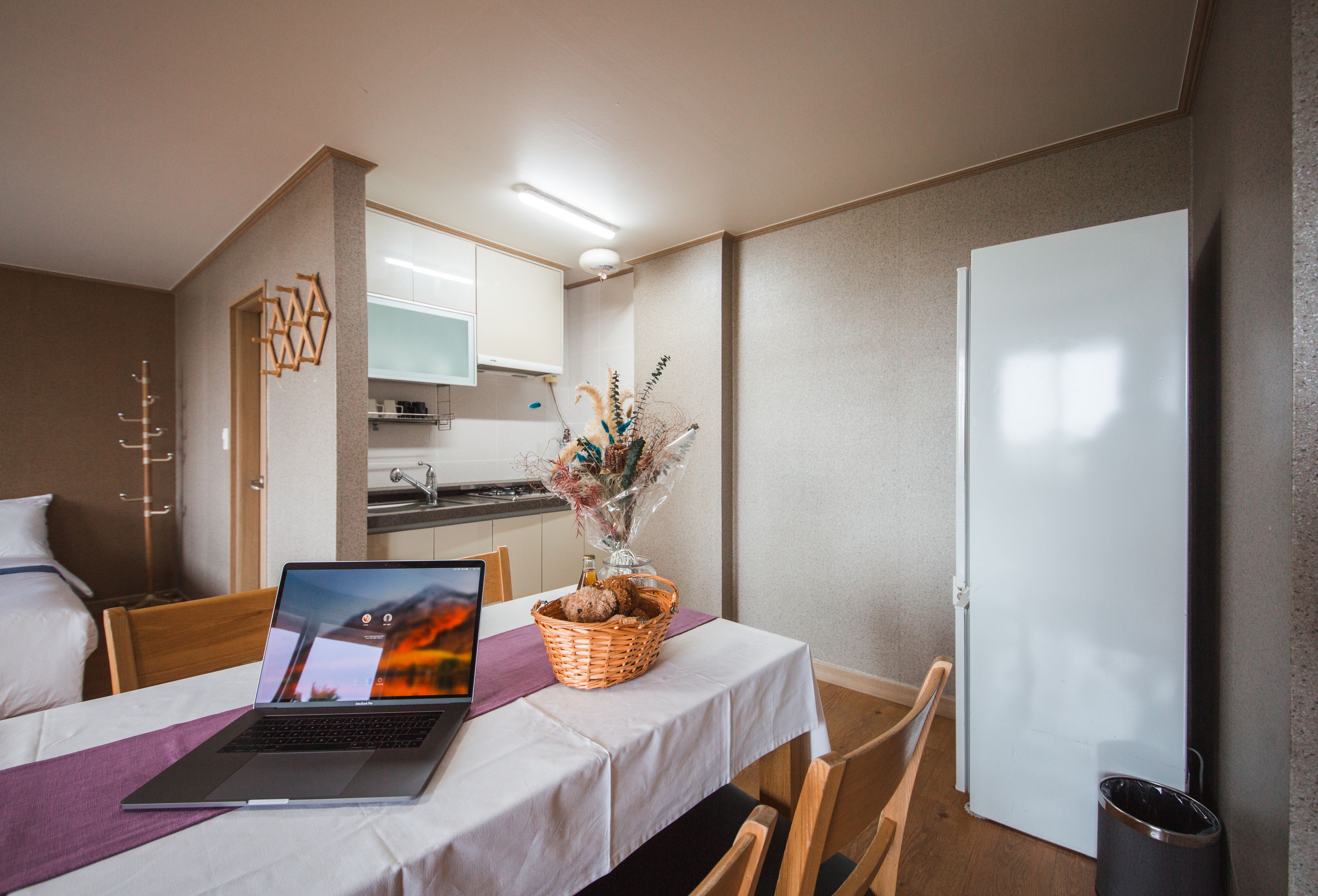 A hotel room with a bed, a dining table with a laptop and a basket of items, a kitchen area, and a white door.