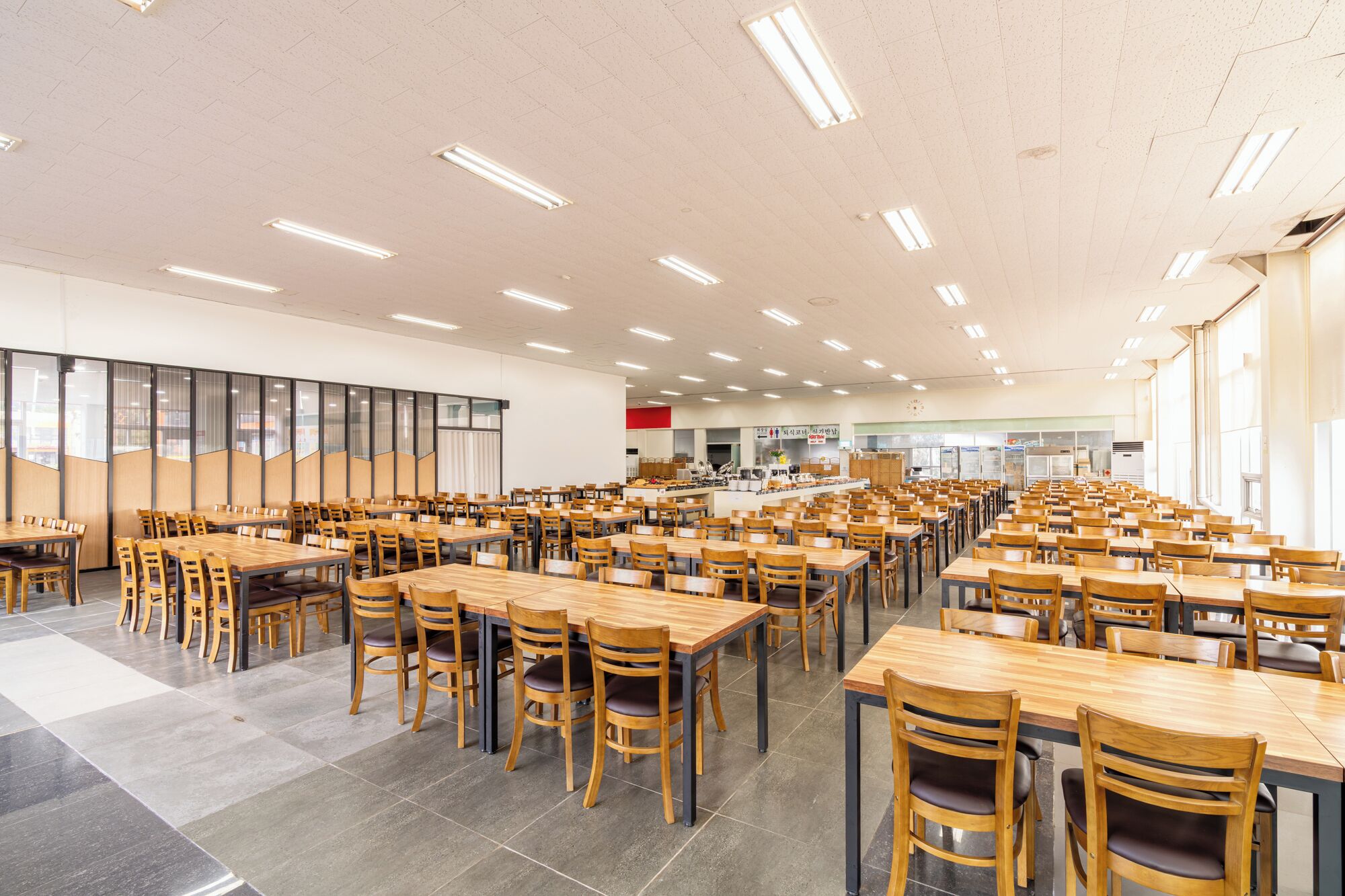 Une salle à manger spacieuse, dotée de nombreuses tables et chaises, d’un coin cuisine et de grandes fenêtres laissant entrer la lumière naturelle.