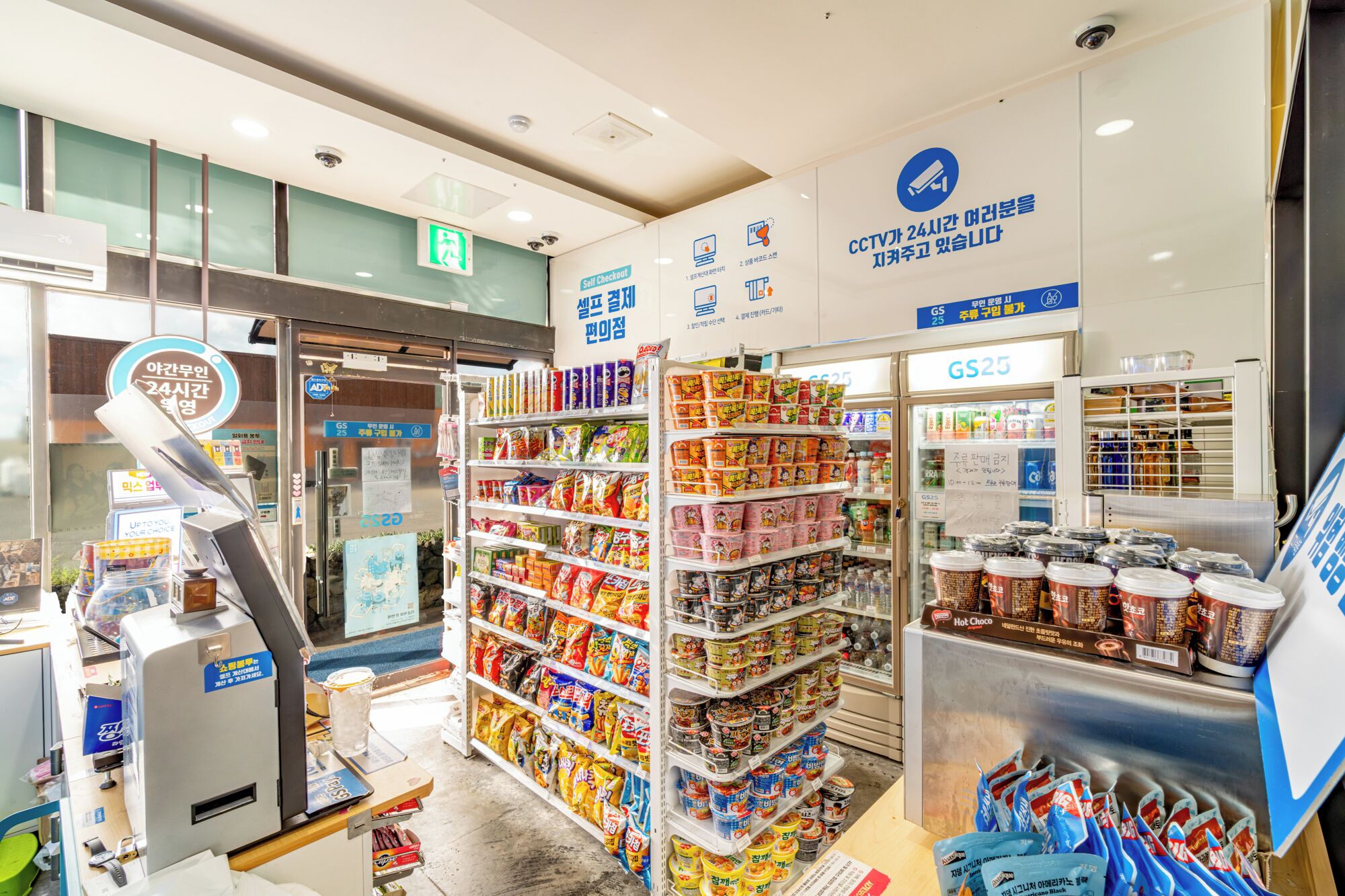 A self-checkout area with a variety of packaged goods on shelves and a refrigerated section with beverages.