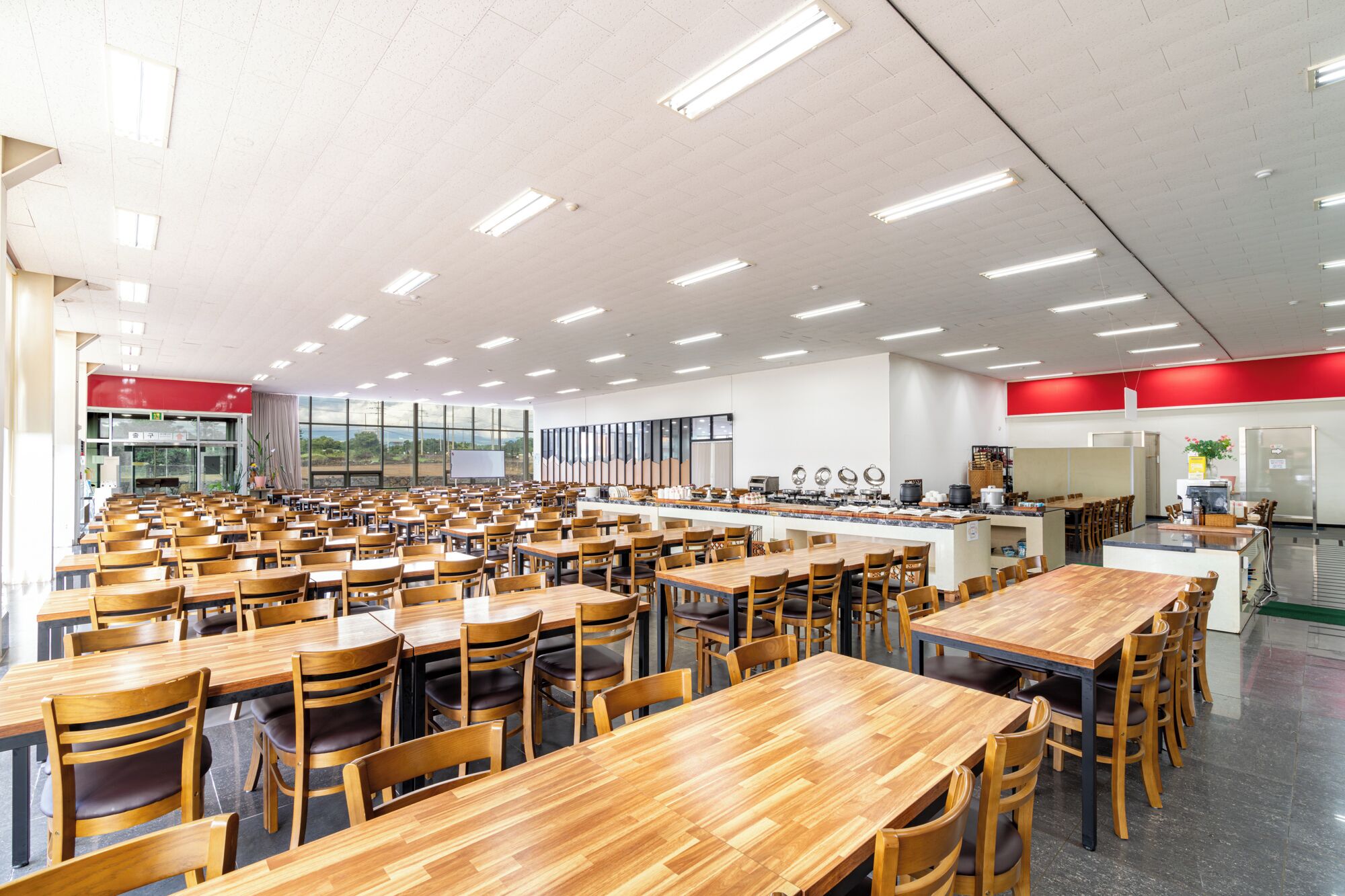 Une cafétéria spacieuse avec des tables et des chaises en bois, un espace cuisine et une grande fenêtre laissant entrer la lumière naturelle.