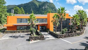 Un edificio moderno con estacionamiento, palmeras y montañas al fondo.
