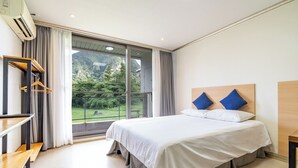 Habitación de hotel con cama, escritorio, silla y un amplio ventanal con vistas a zonas verdes.